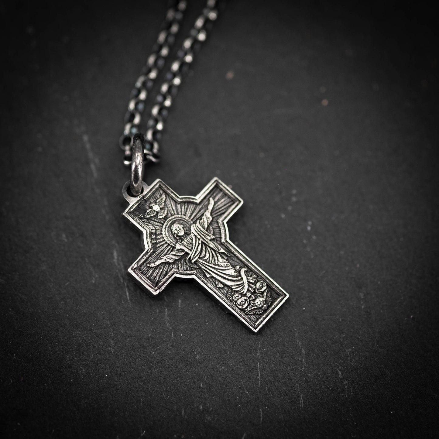 A silver cross pendant with intricate designs, hanging against a dark background.