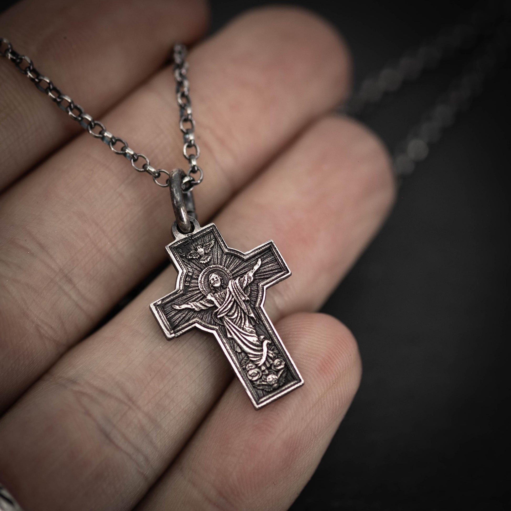 A hand holding a silver cross pendant with a religious symbol.