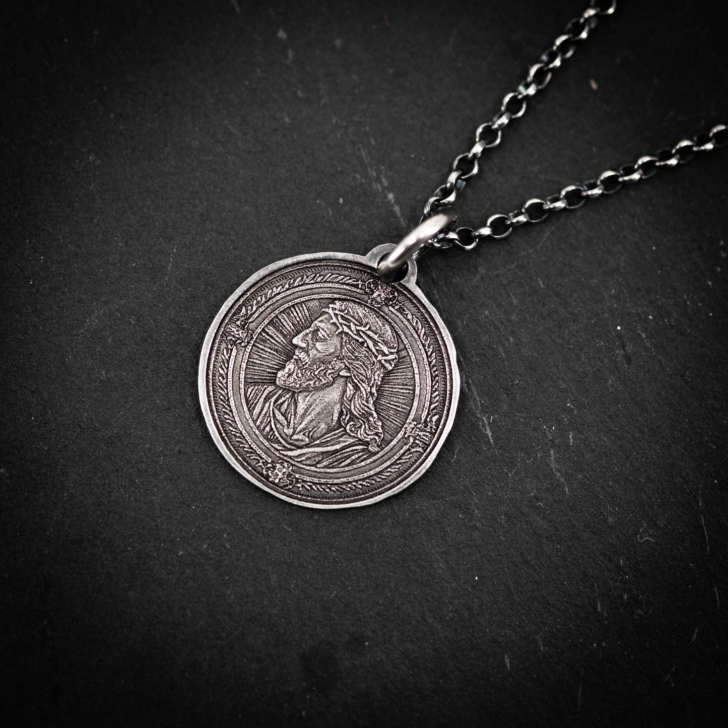 A silver-colored pendant featuring an engraved image of a man's face, possibly depicting Jesus Christ, is displayed on a dark surface.