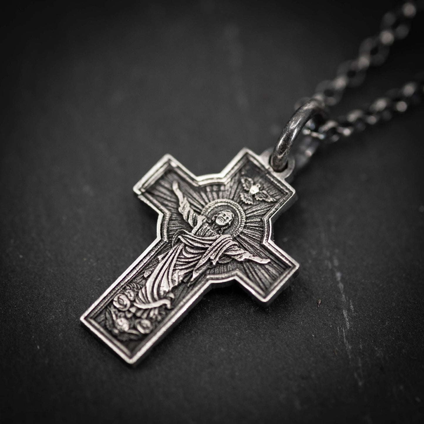 A silver cross pendant with intricate designs and a religious figure on it, hanging from a chain.