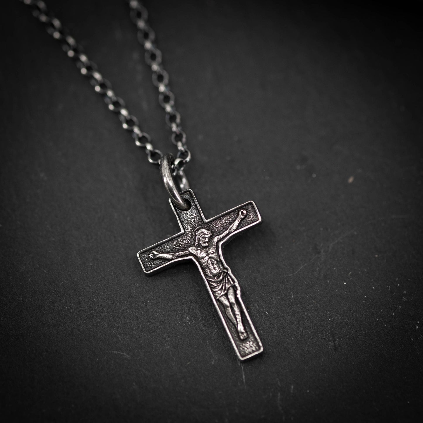 A silver cross pendant with a detailed engraved image of Jesus Christ on it, resting on a dark surface.