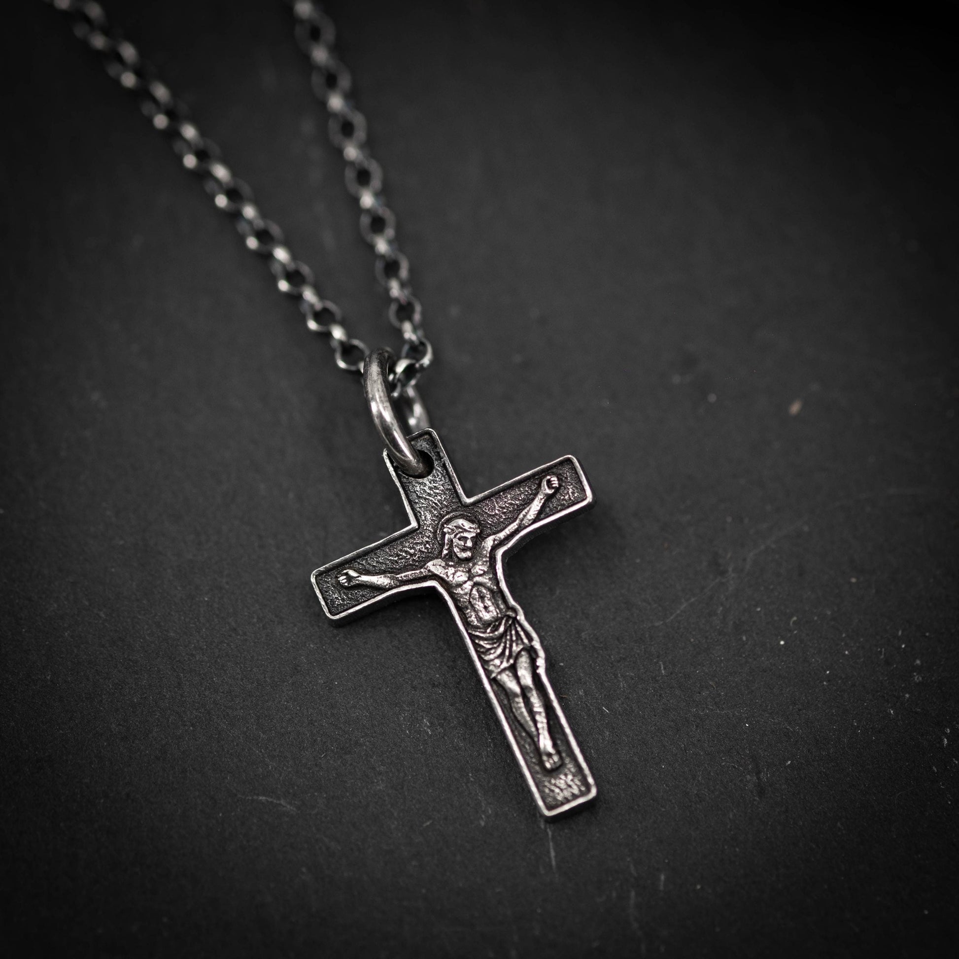 A silver cross pendant with a detailed engraved image of Jesus Christ on it, resting on a dark surface.