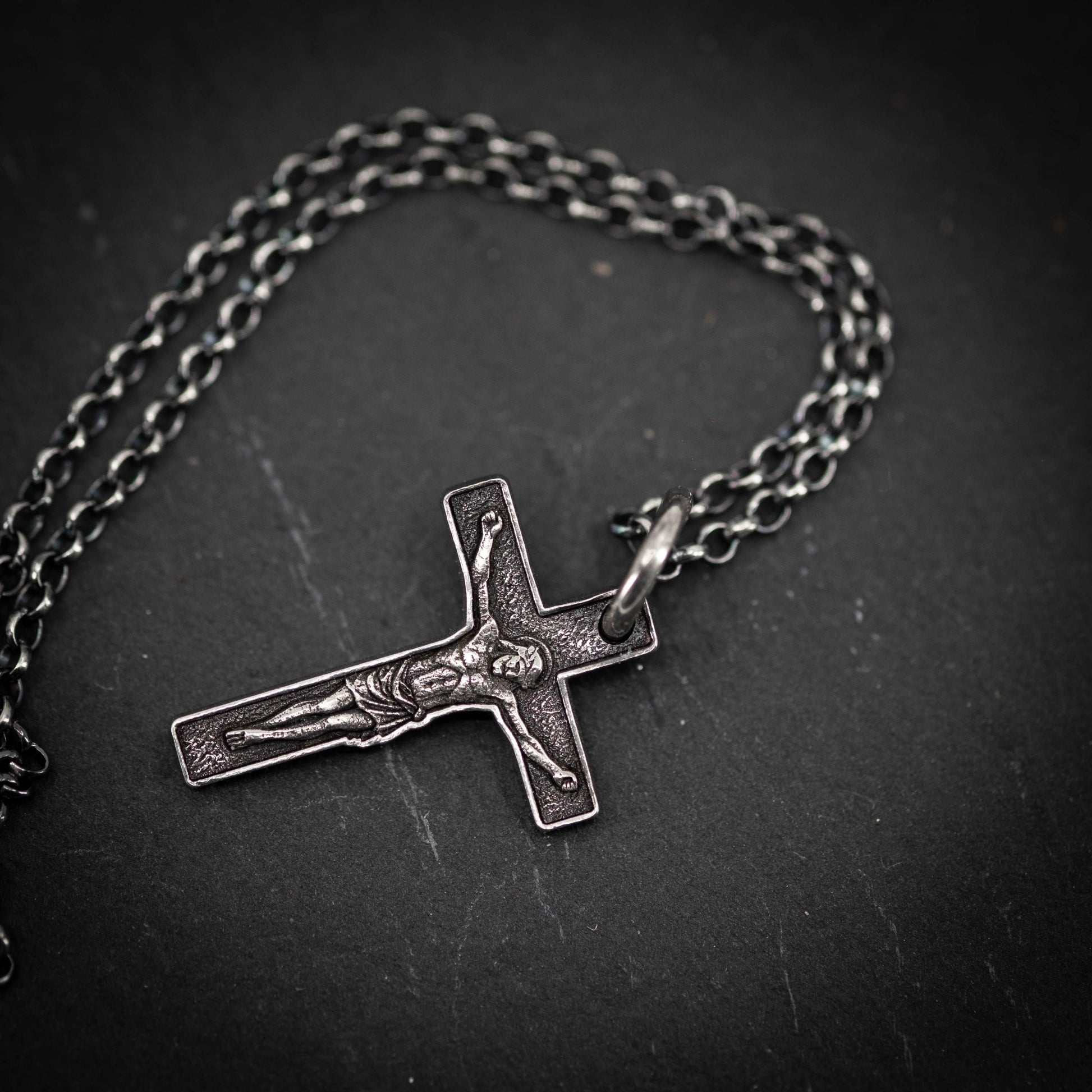 A silver cross pendant with a crucifix design is shown against a dark background.