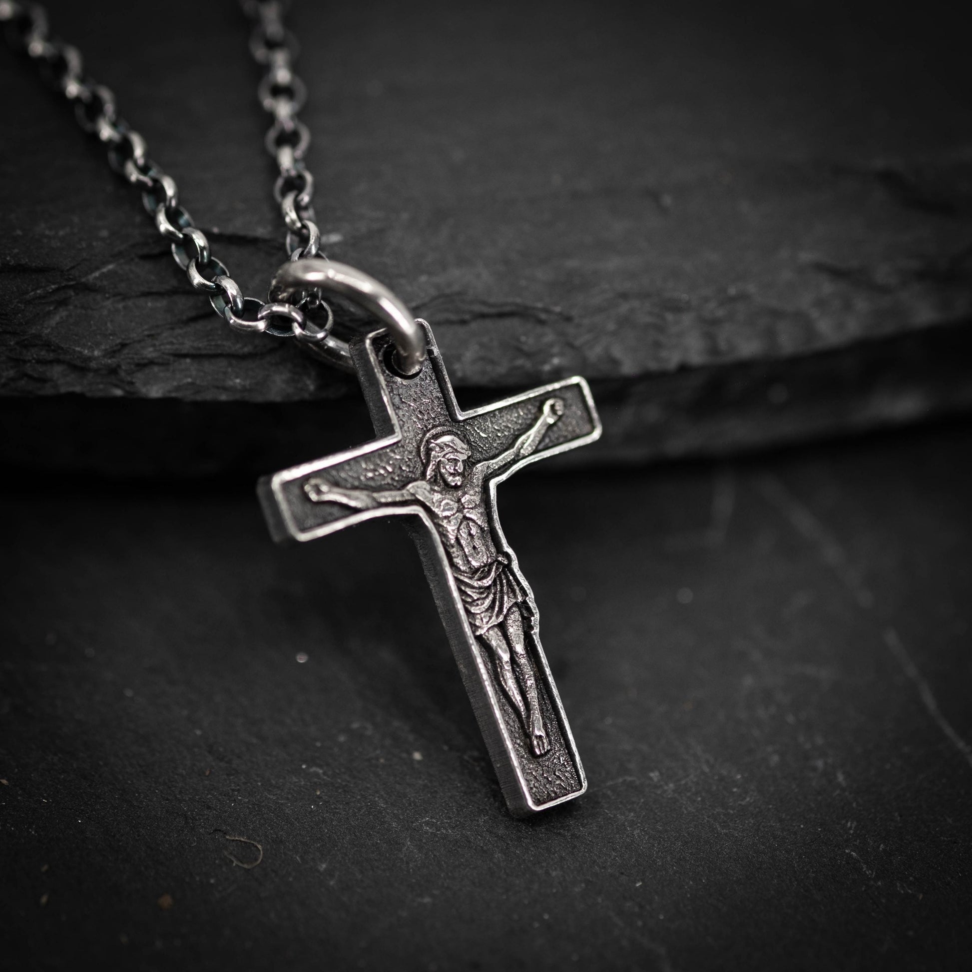 A silver cross pendant with a religious figure engraved on it, hanging on a chain against a dark background.