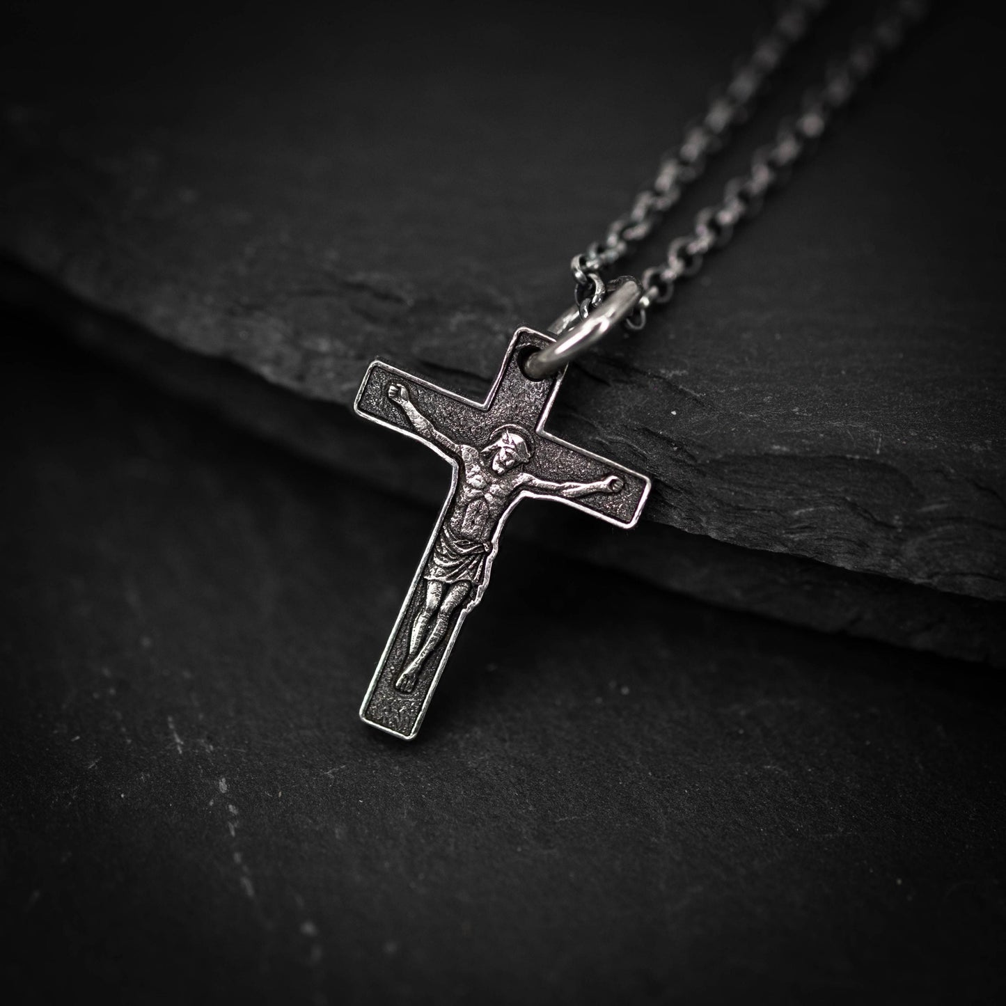 A silver cross pendant with a detailed engraved image of Jesus Christ on it, hanging on a chain against a dark background.