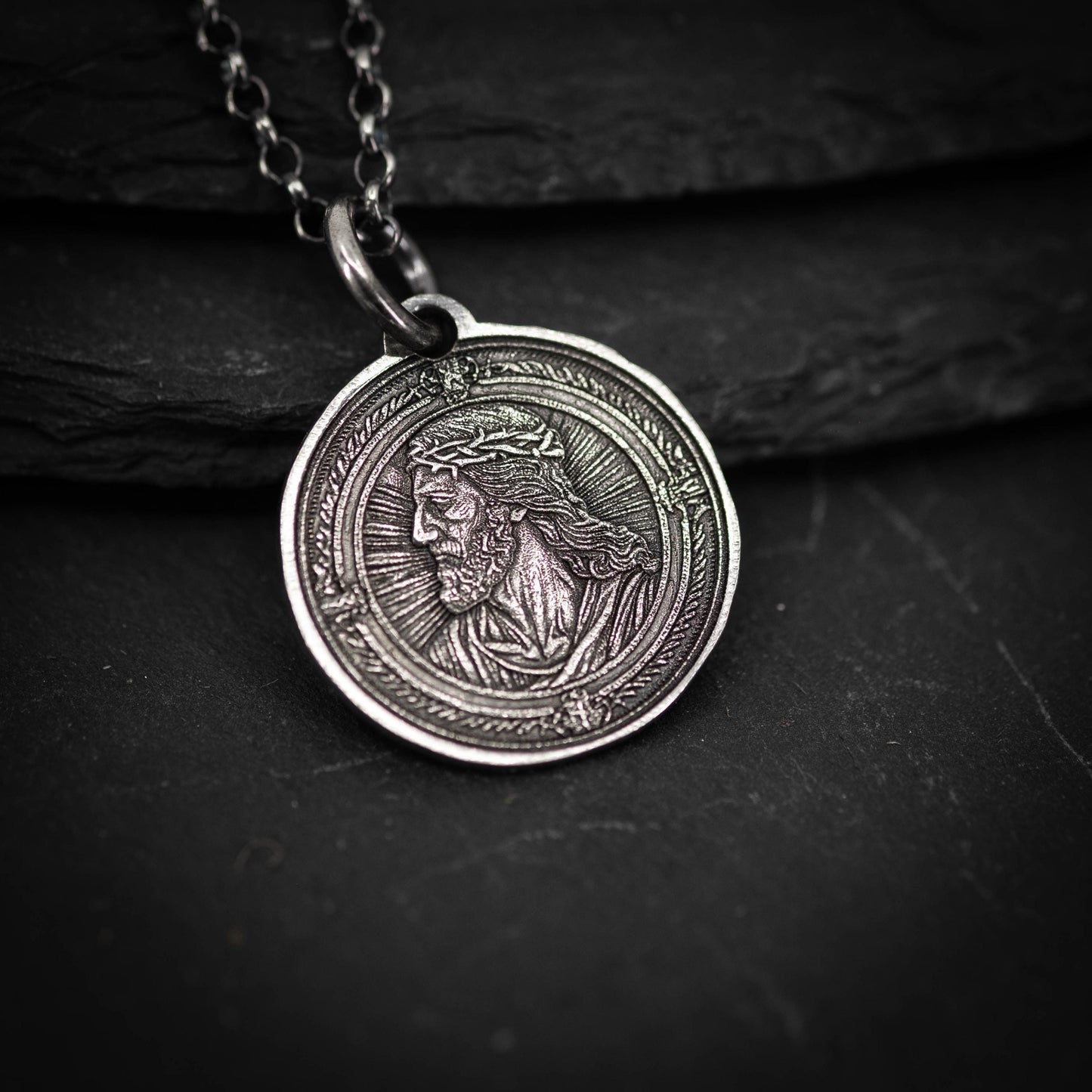 A silver-colored pendant featuring an engraved image of a man's face, possibly a religious figure, is displayed on a dark surface.