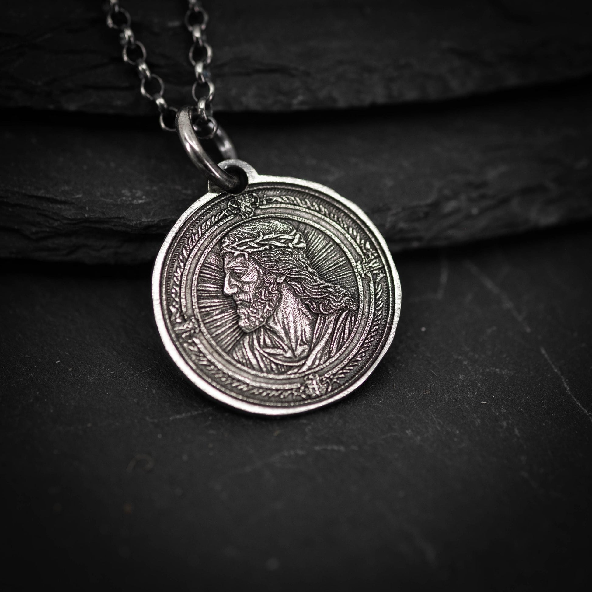 A silver-colored pendant featuring an engraved image of a man's face, possibly a religious figure, is displayed on a dark surface.