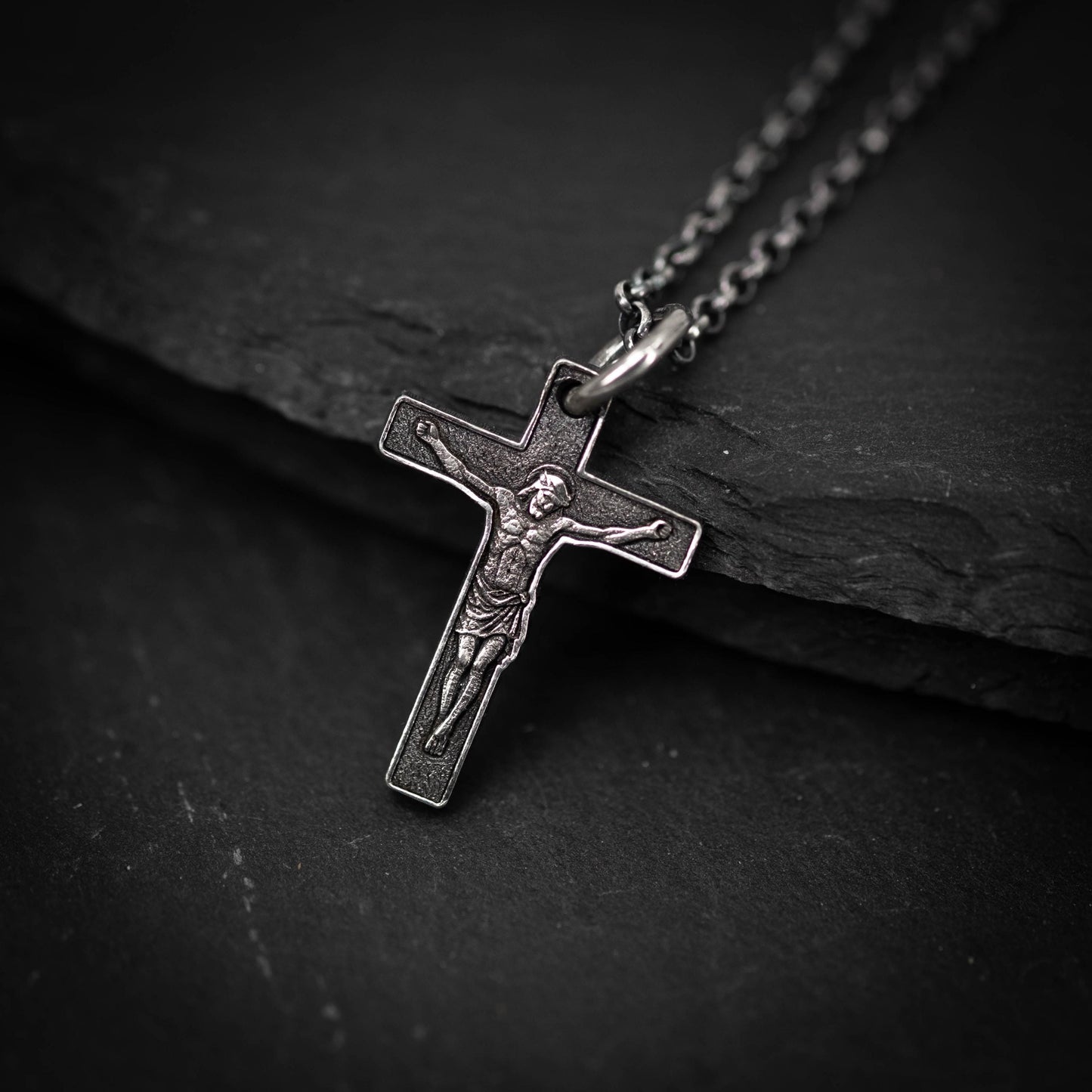 A silver cross pendant with a detailed engraved image of Jesus Christ on it, hanging on a chain against a dark background.