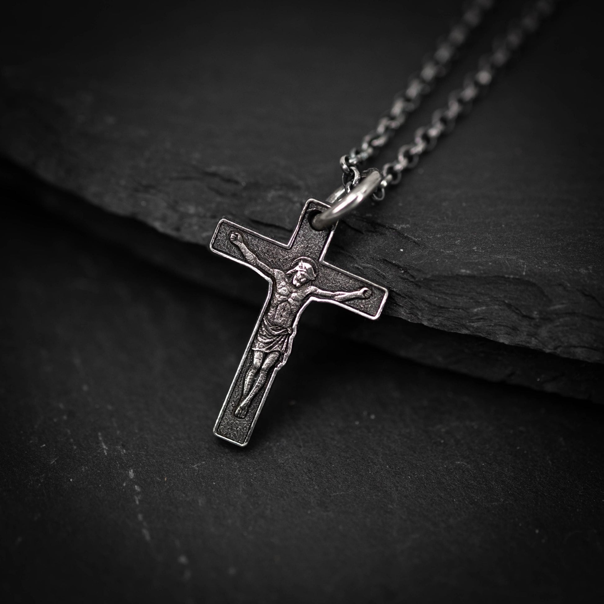 A silver cross pendant with a detailed engraved image of Jesus Christ on it, hanging on a chain against a dark background.