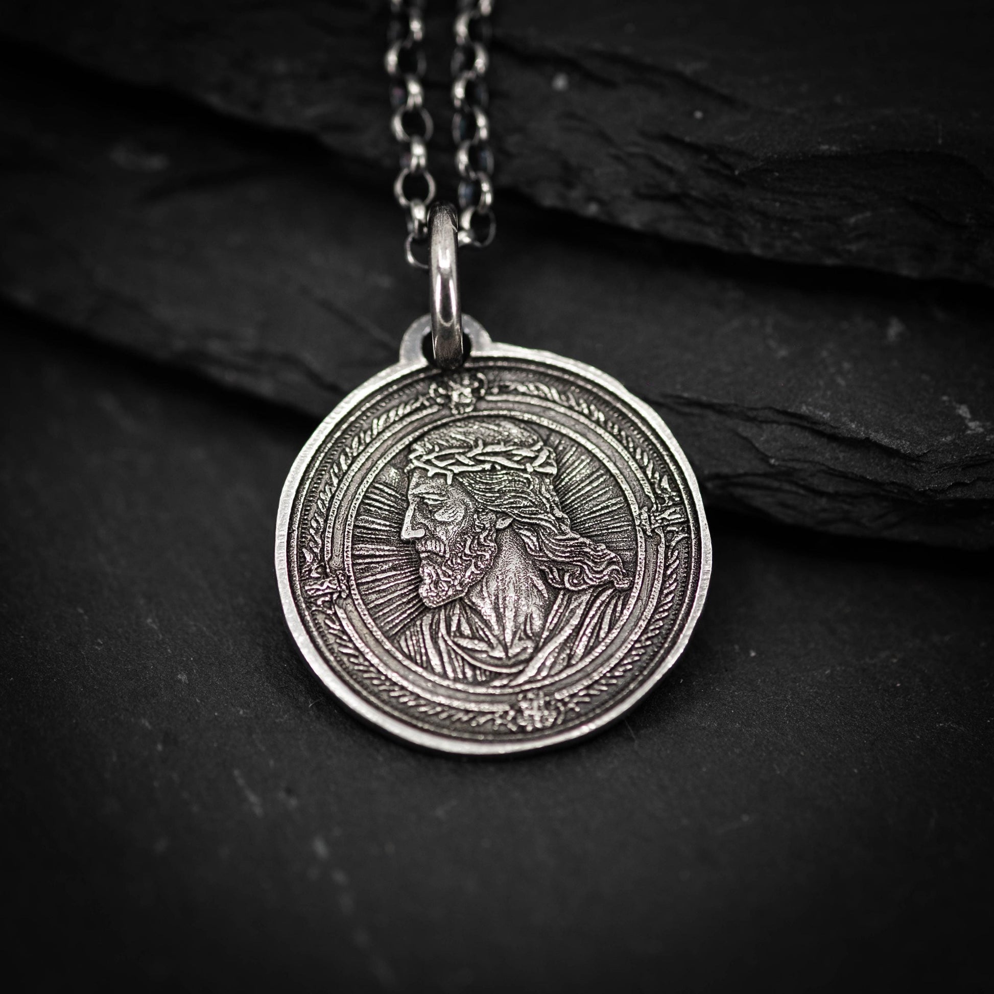 A silver-colored pendant featuring a detailed engraved image of a man's face, possibly depicting Jesus Christ, is displayed against a dark background.
