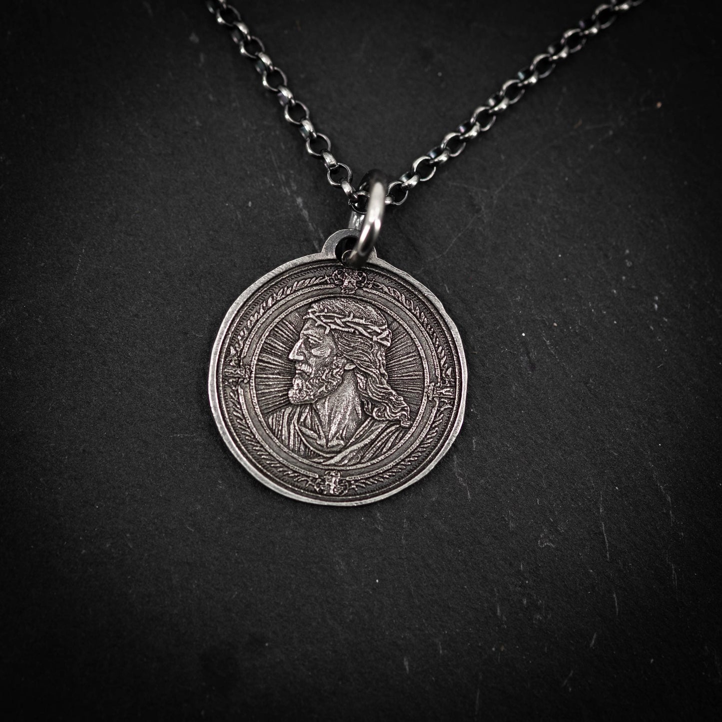 A silver-colored pendant featuring an engraved image of a man's profile, possibly a religious figure.